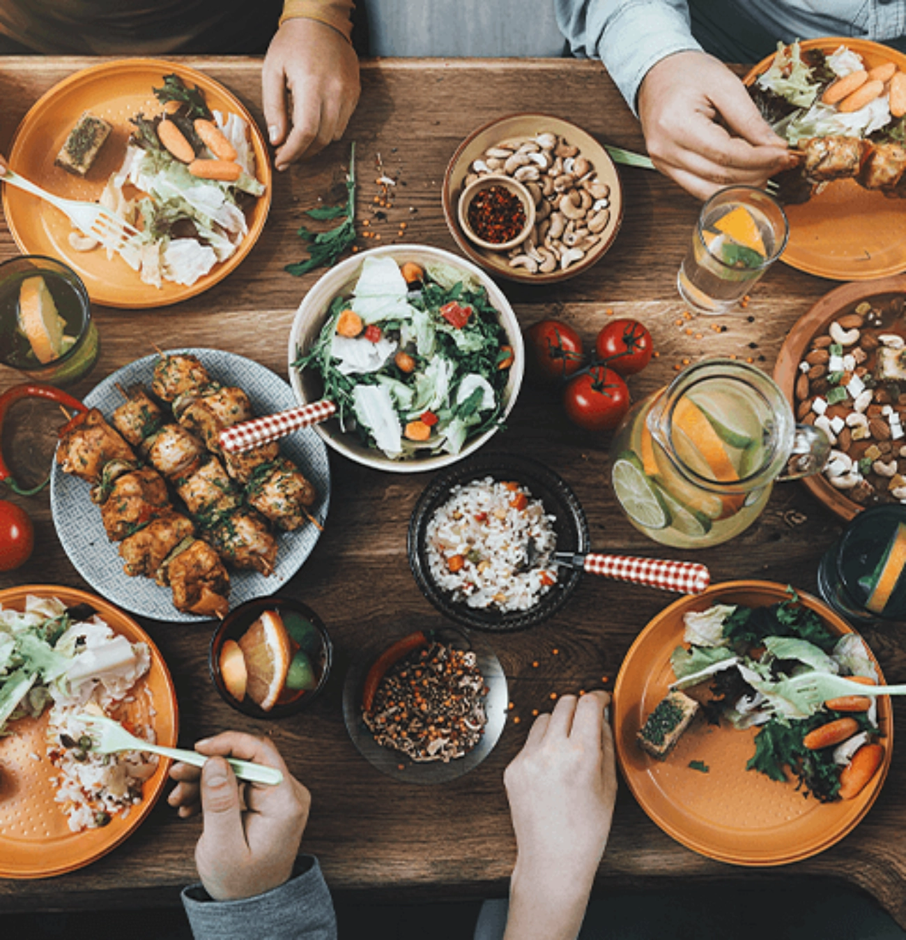 Fotografía de una mesa con diferentes platillos, condimentos y bebidas. Se ven los brazos de cuatro personas comiendo.